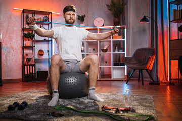 Focused middle aged man training arms with resistance band while sitting on fitball at living room. Caucasian male person spending evening time for training at home. Healthy lifestyles.