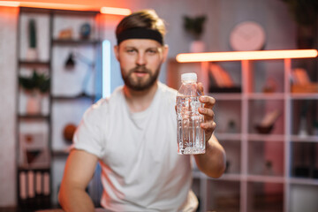 Blurred view of handsome in good mood smiling sporty young bearded man in training clothes, showing a bottle with water to camera when doing home workout in the evening. Focus on the bottle