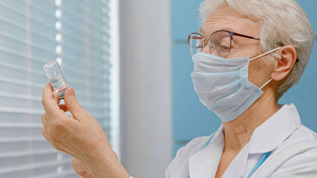Concentrated lady doctor with protective mask fills syringe with vaccine from small vial near window in hospital