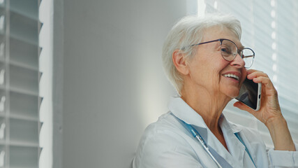 Positive grey haired mature lady doctor with glasses talks on mobile phone with family member near window