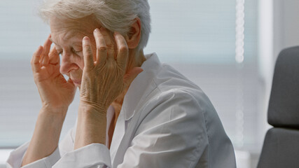 Tired Senior female doctor in coat massages temples suffering from headache in comfortable armchair in clinic office