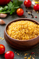 Dry uncooked bulgur in a wooden bowl on a dark background. Bulgur, tomatoes, olive oil, parsley, and spices.