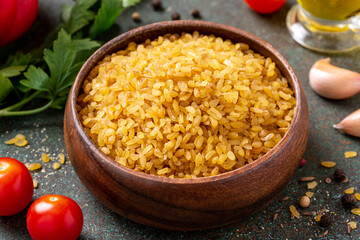 Dry uncooked bulgur in a wooden bowl on a dark background. Bulgur, tomatoes, olive oil, parsley, and spices.