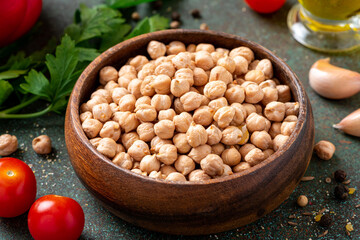 Dry uncooked chickpeas in a wooden bowl and ingredients for cooking on a dark background.