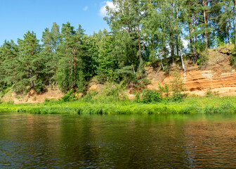 summer landscape from the river, with red, sandstone cliffs on the shore, reflection of trees and shore in the water, Salaca River, Red Cliffs, Latvia