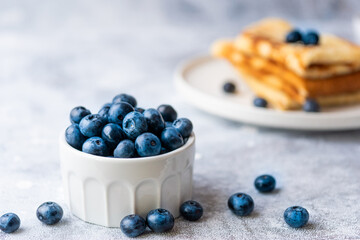 Blueberry sweet summer plant in a white ceramic bowl with pancakes on background in a plate. High key food photography. Sweet breakfast meal.