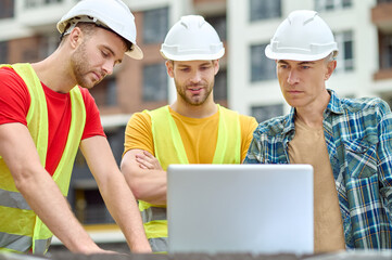 Three concentrated men in protective helmets standing before a laptop