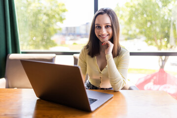 Portrait of beautiful smiling young woman sitting at work station and typing on laptop