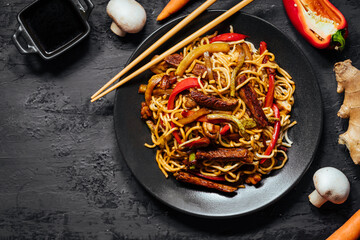Wok with turkey meat, noodles, paprika, mushrooms and carrot served on a black background with chopsticks