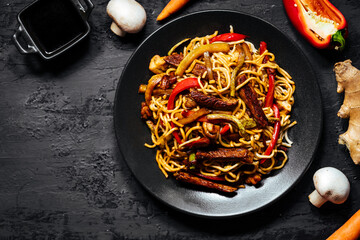 Wok with turkey meat, noodles, paprika, mushrooms and carrot served on a black background with chopsticks