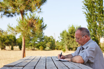 old man writing his memoirs in a blank notepad on a picnic table