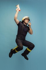 One young male fireman in uniform jumping with loudspeaker and walkie-talkie over blue background - obrazy, fototapety, plakaty