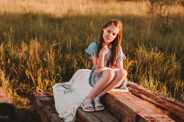 Naklejka na ściany i meble Preteen caucasian girl with long brown hairs wearing blue eco cotton dress sitting down in summer meadow and dreaming. Little girl deep in thoughts.