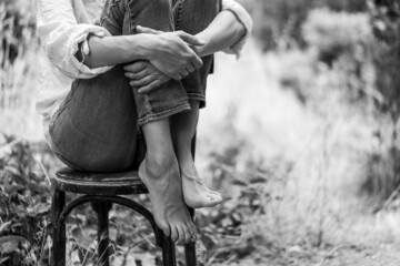 Una Chica joven sentada en una silla en el bosque, con los brazos rodeando sus piernas dobladas y los pies descalzos. En blanco y negro.