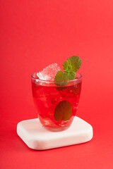 Glass of red refreshing drink with mint on red background. Colorful summer non-alcoholic refreshing drink with ice or fruit iced tea, close-up