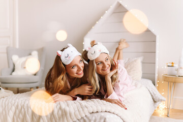 Obraz na płótnie Canvas Pretty two young women in sleep masks laughing relaxing lying on the bed in a cozy decorated bedroom at home during the Christmas holidays, selective focus