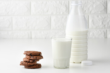a full glass cup of milk and a clear plastic bottle with milk, next to a stack of round chocolate chip cookies on a white table