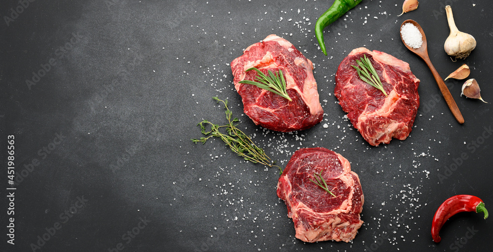 Wall mural raw piece of beef ribeye with rosemary, thyme on a black table, top view