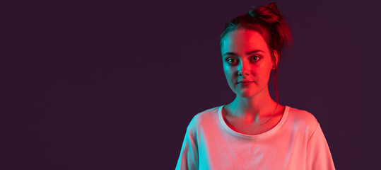Close up portrait of sweet, pretty romantic girl on dark purple studio background in red neon...