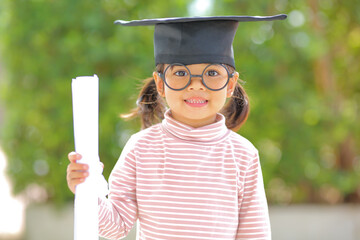 A little girl asia looks happy while celebrating his graduation and holding a diploma and graduation cap  which increases the development and enhances outside the classroom learning skills concept.