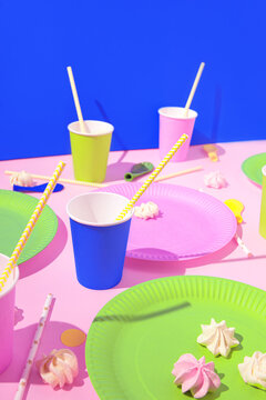 Creative composition made of colorful paper glasses with straws, dishes, meringues and balloons on bright pink and blue background. Festive concept. Birthday and party theme.