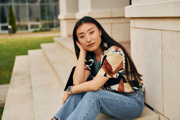 Sits on the stairs of building. Young asian woman is outdoors at daytime