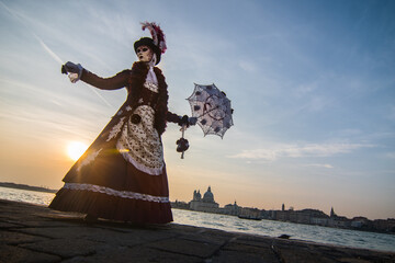 Female wearing mask and carnival costume posing by the water with an umbrella in Venice, Italy