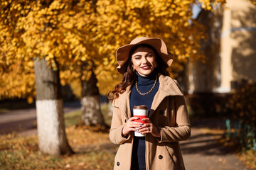Stylish woman dressed in beige coat and wide brimmed hat holds a thermocup, outdoors in autumn.