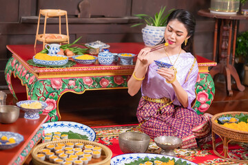 A Thai Asian woman wearing a traditional Thai dress from the Ayutthaya period. She is sitting in the kitchen eating Thai desserts. - obrazy, fototapety, plakaty