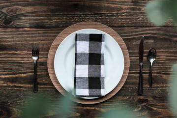 Flat lay of a place setting with buffalo plaid napkin, silverware and charger over a rustic wood table looking through the greenery of a houseplant. Selective focus with blurred foreground.