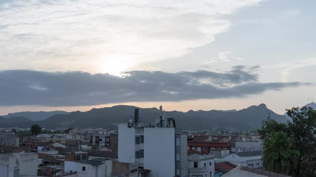 Time lapse video of a sunset with the sun behind some clouds and then setting through the mountains, from the terrace of a building and an urban background of houses and buildings.