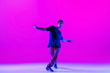 Young stylish man, hip-hop dancer dancing solo in modern clothes isolated over bright magenta background at dance hall in neon light. - obrazy, fototapety, plakaty