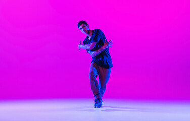 Fototapeta na wymiar Young stylish man, hip-hop dancer dancing solo in modern clothes isolated over bright magenta background at dance hall in neon light.