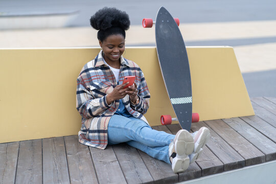 Cheerful black female with skateboard chat on smartphone with smile sit in skate urban park use phone texting outdoors. Happy african american skater girl with longboard check mobile app notifications - Powered by Adobe