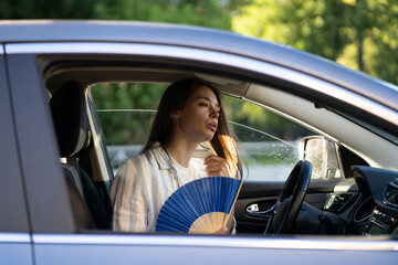 Exhausted young woman driver with hand fan suffering from heat in car, has problem with a non-working air conditioner, try to cool herself on hot sunny day. Summer season concept. 