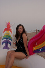 Portrait of young woman enjoying the end of summer drinking a glass of wine while sitting on inflatable unicorn float on the beach sand and smiling to the camera. Summer Girl 2021.