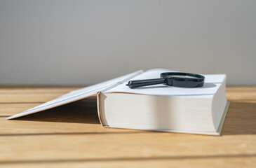 Magnifier with open thick book on wooden desk with daylight.
