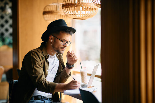 Young man writes in notebook while uses laptop and studying in cafe.