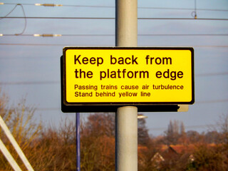Yellow sign warning people to keep back from the edge of a railway station platform when trains are passing.