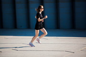 Fototapeta premium a white girl with short blonde hair in black clothes and light sneakers runs against the background of a gray concrete wall. photo motion drive