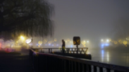 A mysterious figure standing by street lights next to a river on a misty winters night. With a blurred, bokeh, out of focus edit