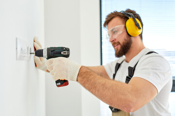 Sockets installation. Caucasian handyman setting new sockets in living room. Home renovation. Technician in overalls, safety eyeglasses and headset is concentrated on work, using electric drill