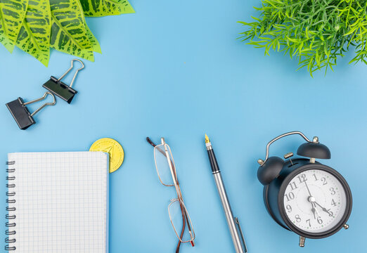 Business conceptual flat lay still life with crypto coin and alarm clock alongside a notepad, reading glasses and pen on a blue background with fresh green plants in the corners