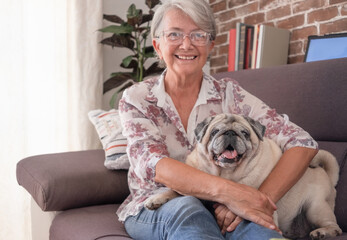 Old pug dog sitting on the sofa at home with a senior woman smiling and hugging