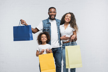 pleased african american family with multicolored shopping bags smiling at camera on grey