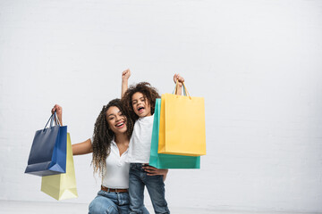 excited african american mom and daughter with multicolored shopping bags on grey