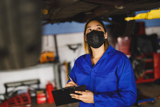 Mixed race female car mechanic wearing face mask and overalls, making notes on clipboard