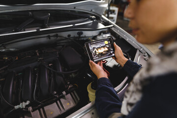 Hands of mixed race female car mechanic wearing overalls, inspecting car, using tablet