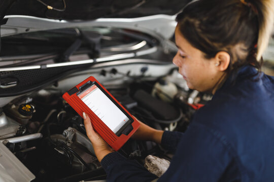 Mixed race female car mechanic wearing overalls, inspecting car and using tablet