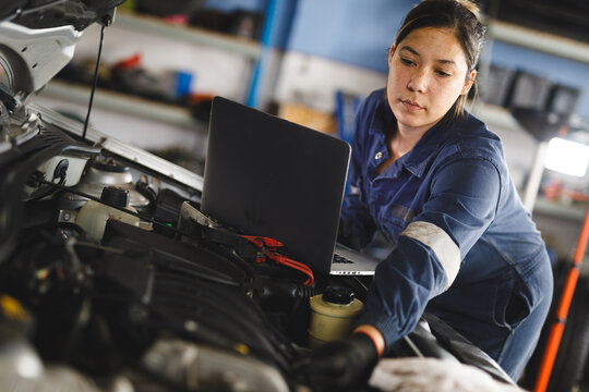 Mixed race female car mechanic wearing overalls, inspecting car, using laptop